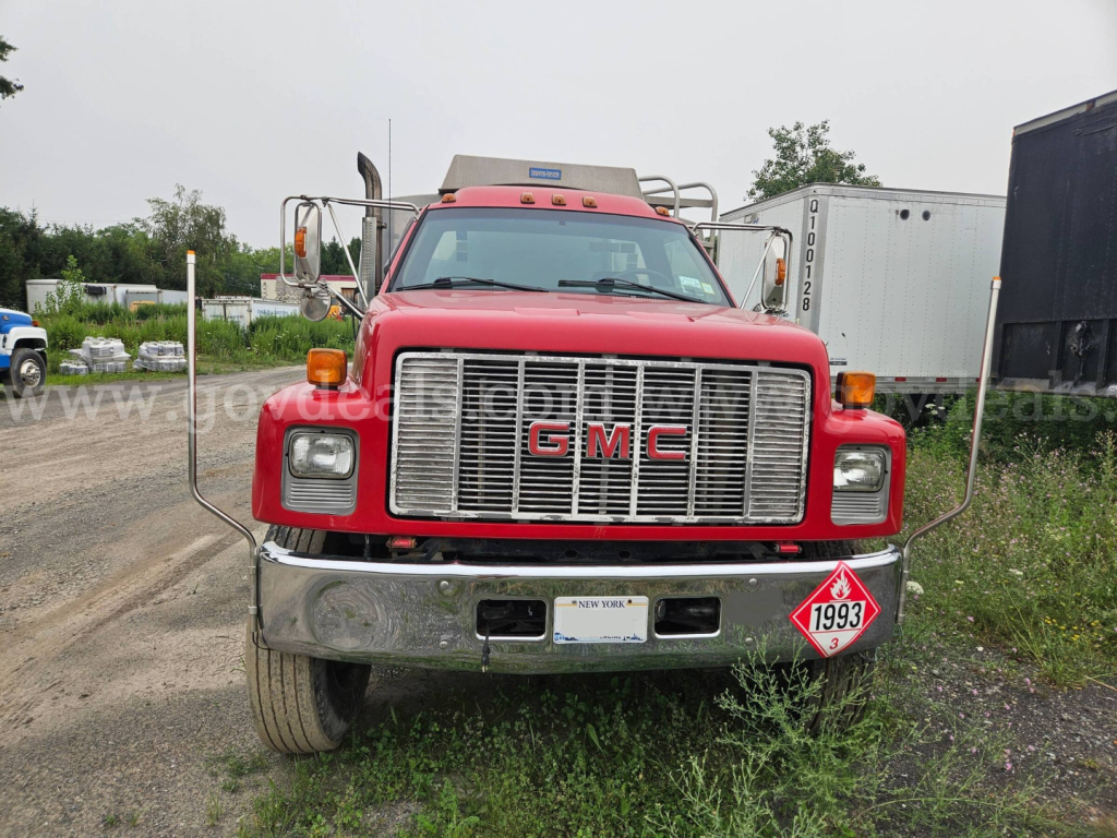 GMC C7500 front view showing chrome grille, bumper, and hazard placard on lower right; red cab with dual mirrors and light bar.