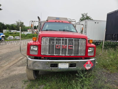 GMC C7500 front view showing chrome grille, bumper, and hazard placard on lower right; red cab with dual mirrors and light bar.