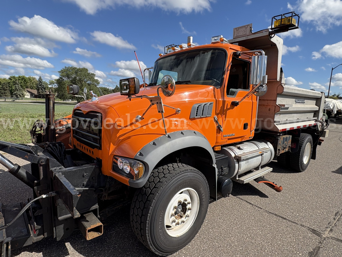 Truck cab of a 2010 Mack GU712 plow truck in orange, side view showing front wheel, windshield, and plow mount.