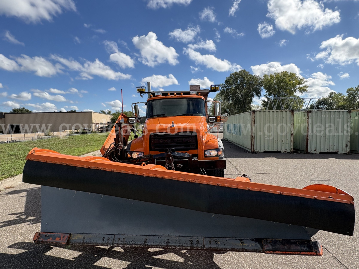 Front view of an orange 2010 Mack GU712 plow truck with a large snow plow blade attached, in an outdoor lot with storage containers and blue sky.