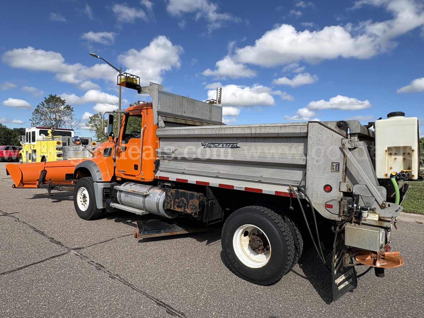 Mack GU712 dump truck plow show­ing side view with orange cab and gray dumping bed, plow attached at front, side steps, wheel, and tailgate device visible.