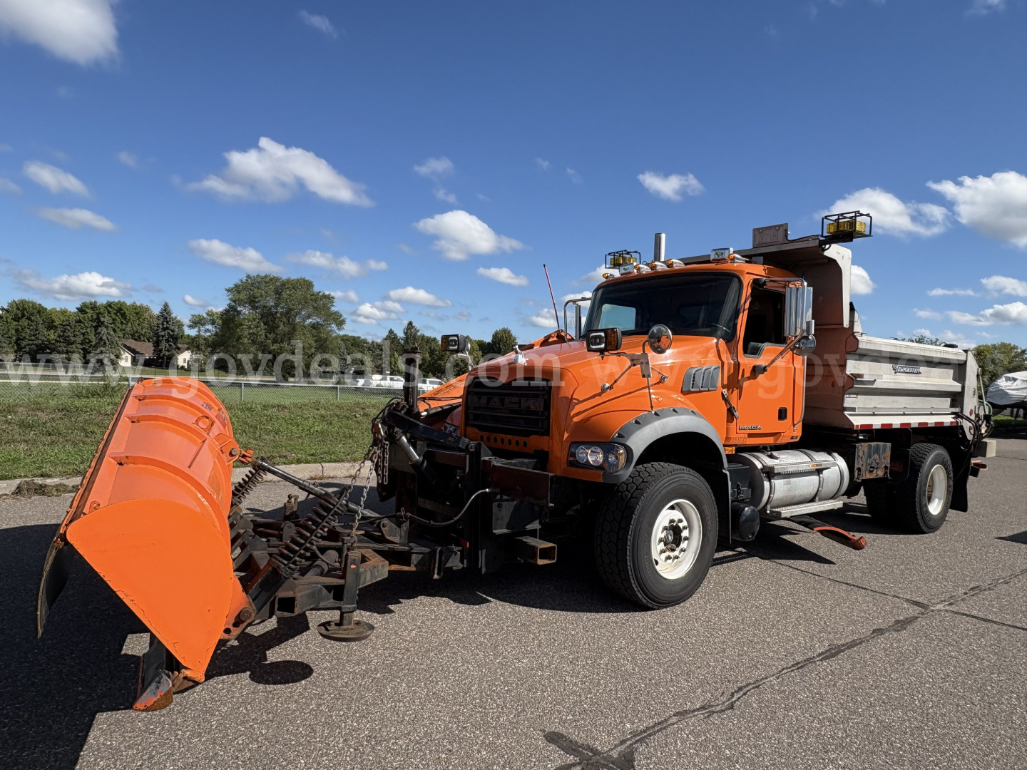 Mack GU712 dump truck with orange plow attachment and front-mounted blade showing in a parking lot.