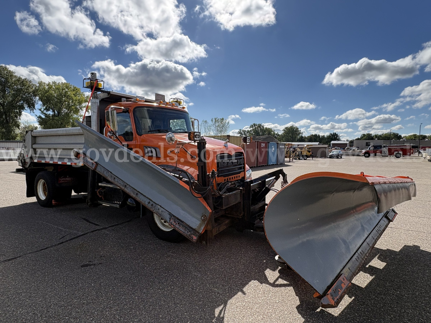 Mack GU712 dump truck with an attached snow plow blade in a parking lot. Side ramps extended, orange cab, gray plow, clear blue sky.