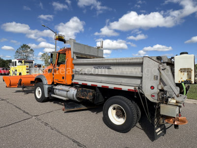 Mack GU712 dump truck plow show­ing side view with orange cab and gray dumping bed, plow attached at front, side steps, wheel, and tailgate device visible.