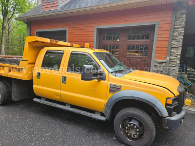 Ford F-450 dump truck in yellow with gray fender flares, side steps, and utility bed parked in front of orange building.