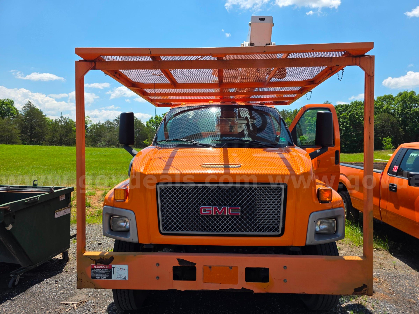 GMC bucket truck front view with orange Altec TA45M attachment on a bright day; grille with GMC badge, large front bumper, raised platform over cab.