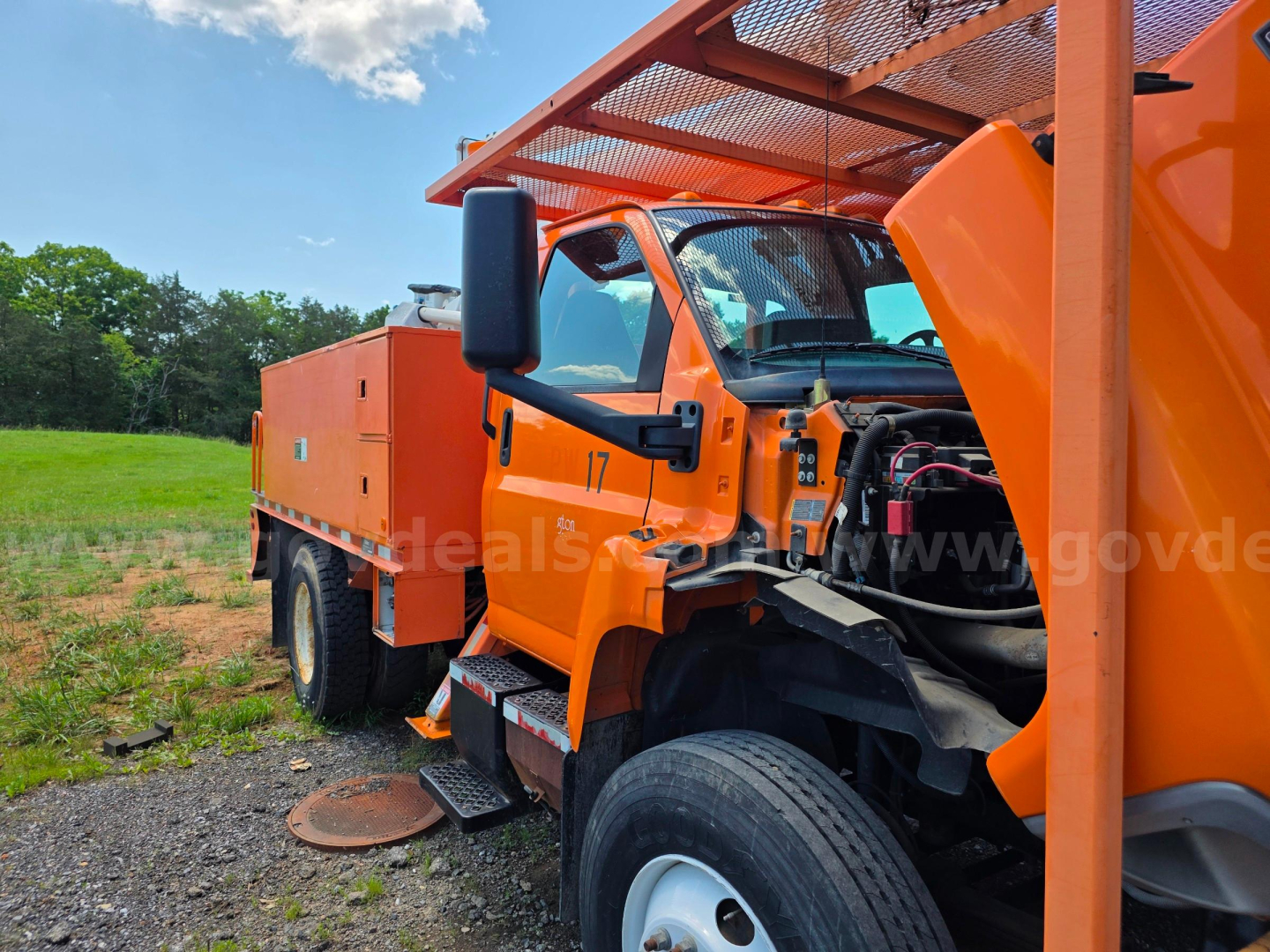 Orange Altec TA45M bucket truck with damaged front left area and exposed engine compartment; wheel and grounding pad on ground; clear blue sky and grassy background.