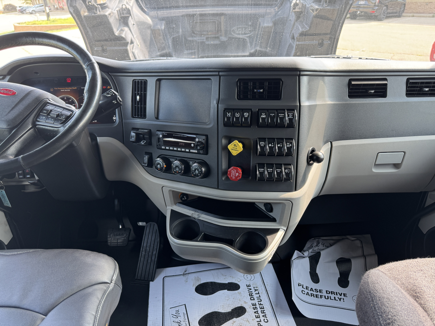 Peterbilt 579 cabin dash and steering wheel showing infotainment cluster and control switches; center console with cup holders and storage; windshield with hood open.