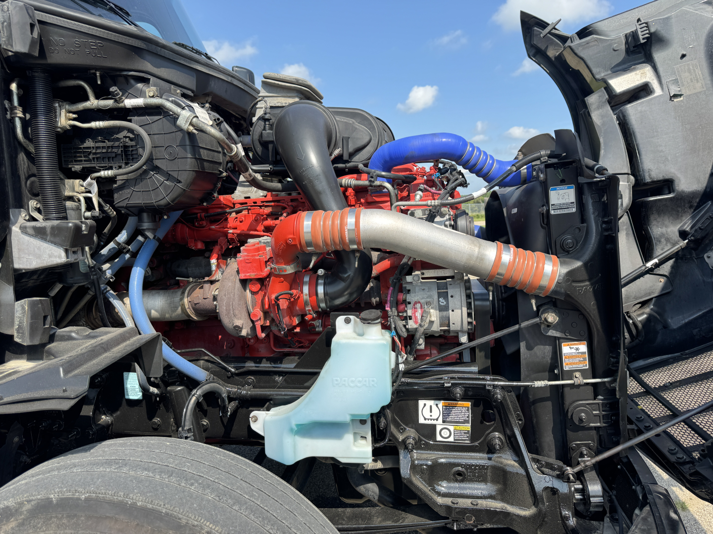 Peterbilt 579 engine bay showing blue coolant reservoir, orange and blue hoses, red engine block, and surrounding components.