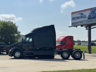 Peterbilt 579 truck tractor in profile view with a trailing trailer chassis, black exterior, sleeper cab, highway lot, signboard in background.