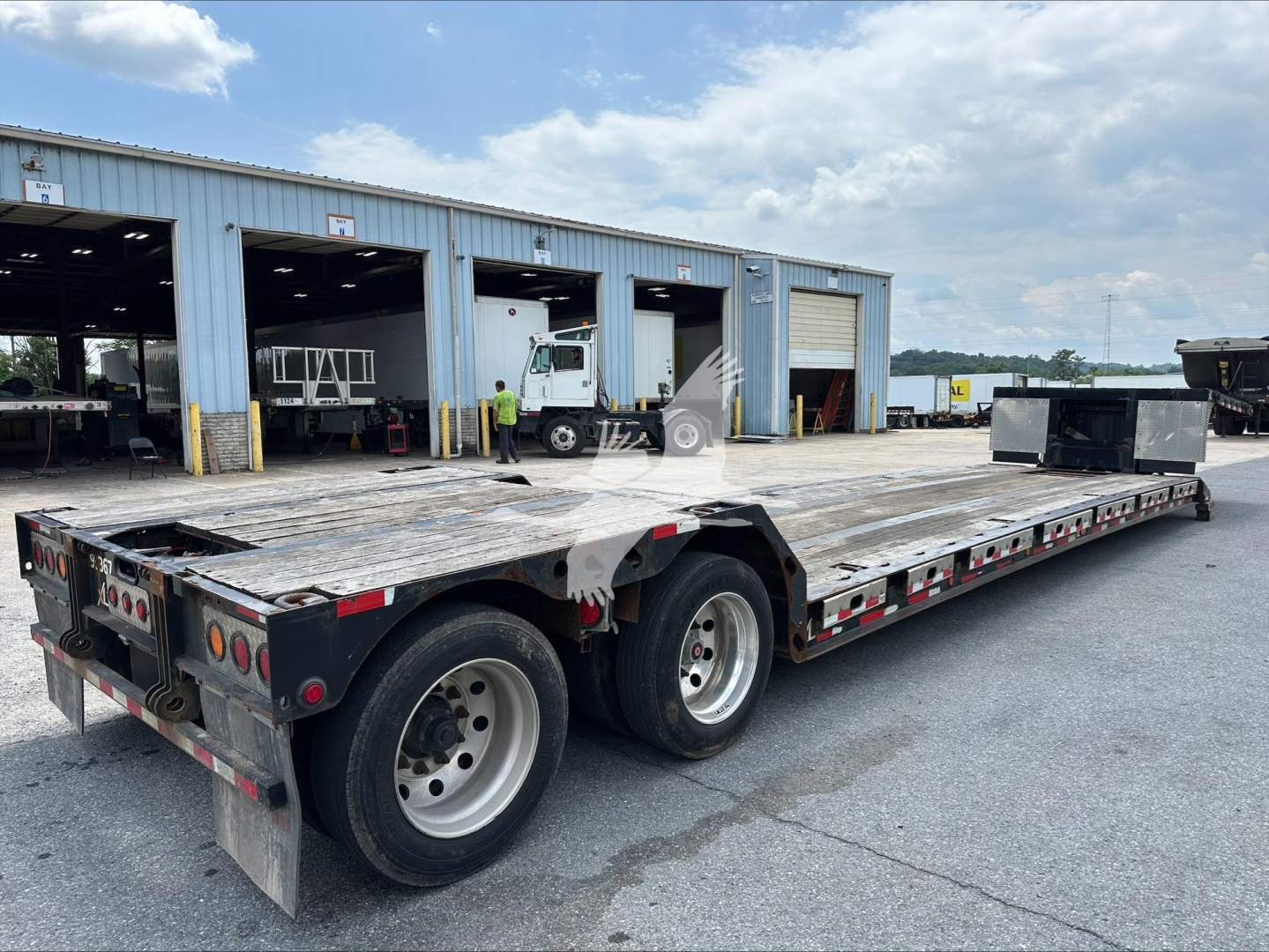 Flatbed trailer with dual rear axles in an open lot, showing a long aluminum deck and black undercarriage; background has warehouse buildings and a few workers.
