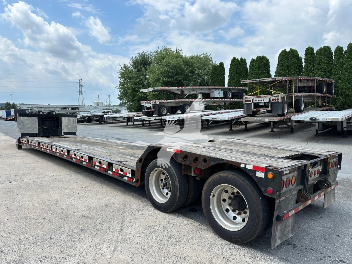Flatbed trailer with dual rear axles and long metal deck, storage boxes on front, multiple trailers in background, no visible text on the side.