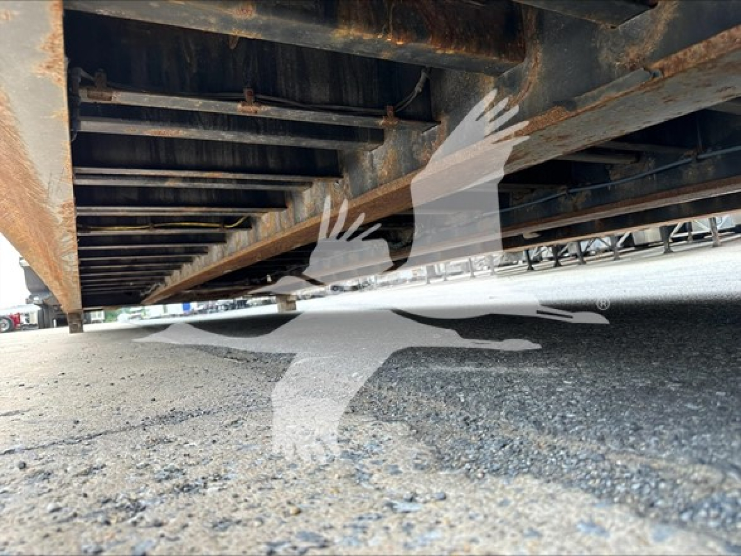 Trailer underside with frame rails and crossmembers visible; rusting metal and gravel ground.