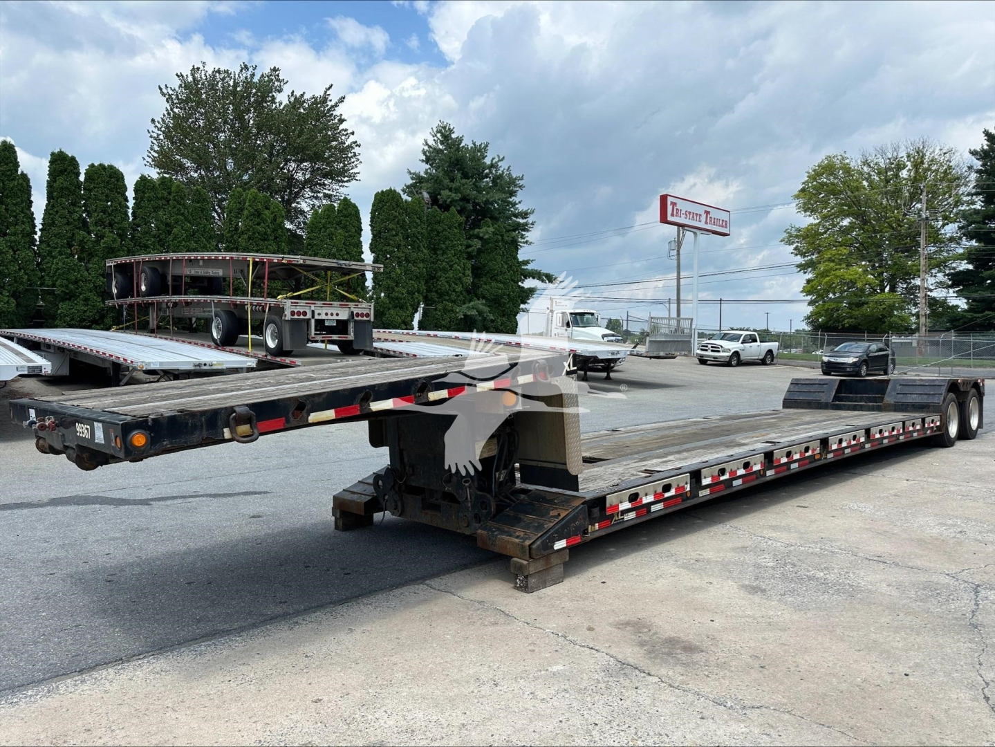 Low-angle view of a long flatbed trailer with extendable double drop deck in a dealership lot. In the background are trees, a few vehicles, and a sign. The trailer shows a metal surface with tie-down points and reflective tape along the sid