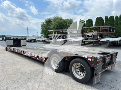 Flatbed trailer with dual rear axles and metal decking, long low-profile trailer in an outdoor lot with other trailers in background.