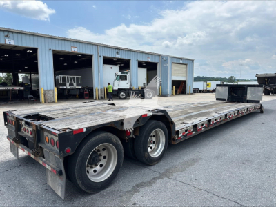 Flatbed trailer with dual rear axles in an open lot, showing a long aluminum deck and black undercarriage; background has warehouse buildings and a few workers.