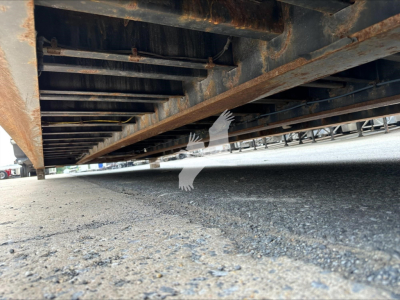 Trailer undercarriage showing steel frame and rust-wrapped beams of a double drop expandable trailer; asphalt ground visible beneath.