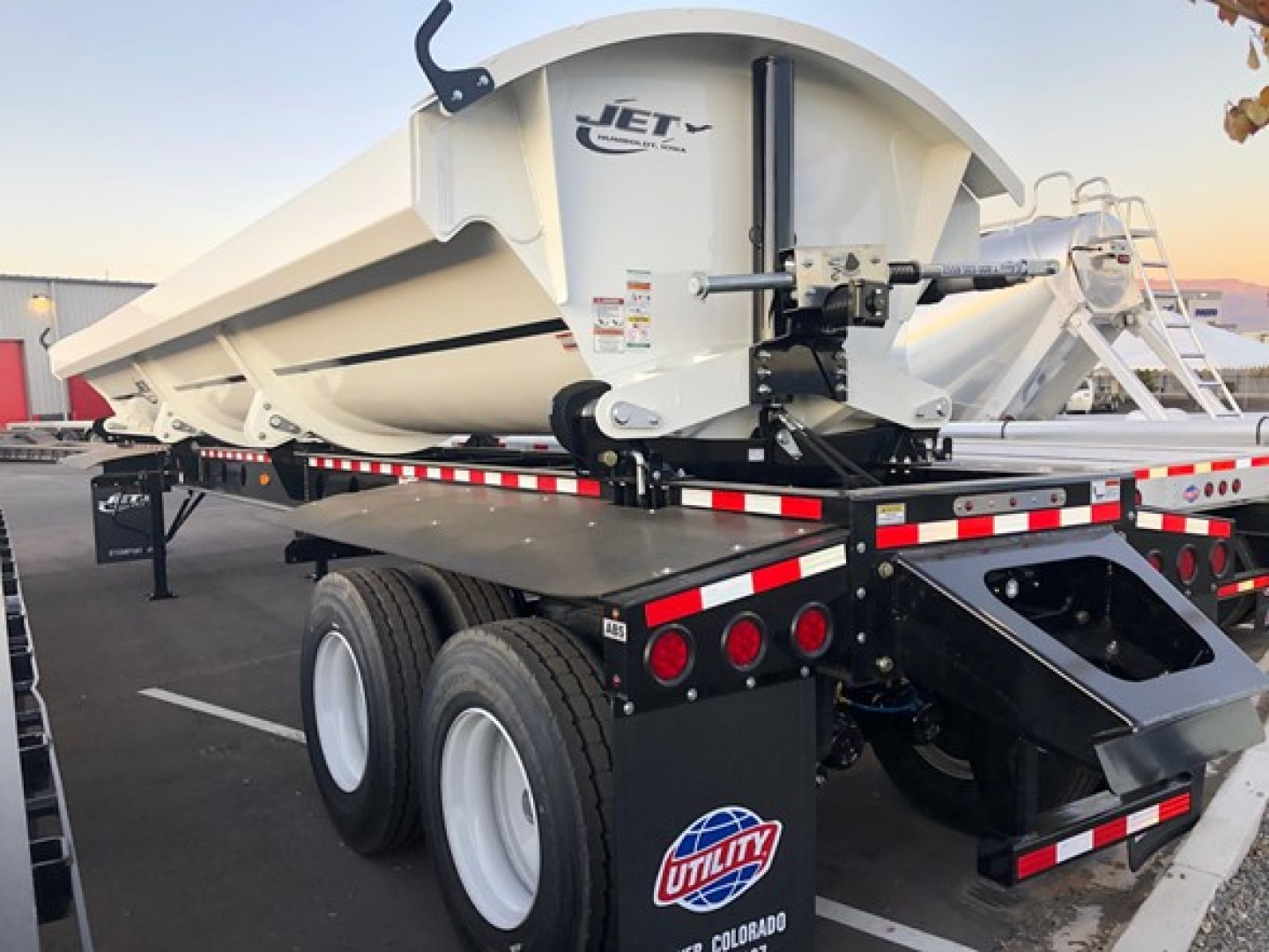 Jet trailer side dump with white hopper mounted on a tri-axle trailer. visible text includes 'JET' on the hopper and warning decals on the side near the hitch. Aluminum ladder/rack and reflective safety tape along the rails.