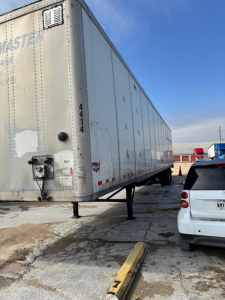 Rear-left view of a white Wabash trailer with a raised landing gear and a black door handle, showing some faded markings and a small vent; a white SUV is parked nearby.
