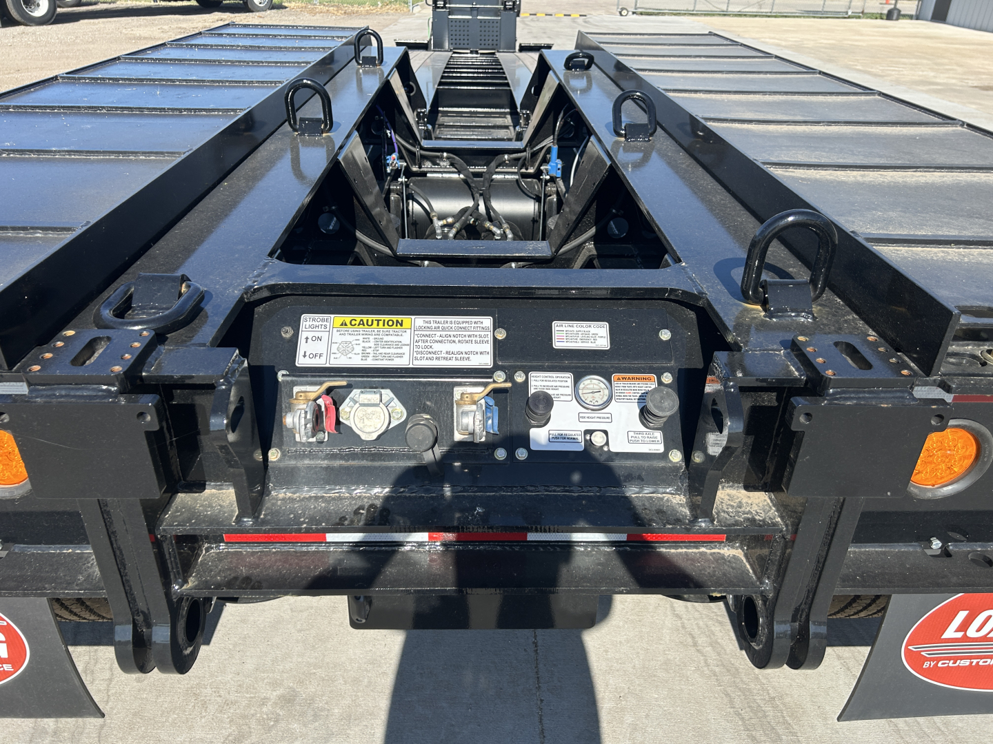 Trailer hitch area of Load King lowboy trailer showing control panel with warning label, latching hardware, and hydraulic components.