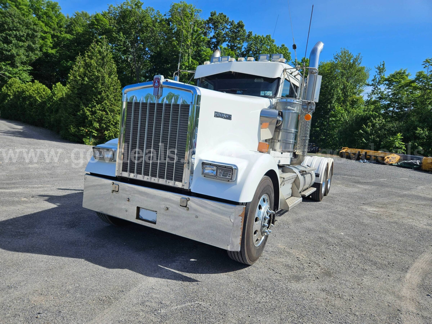 Kenworth W900L semi truck front-left view showing grille, bumper, headlights, and exhaust stacks in a truck yard.