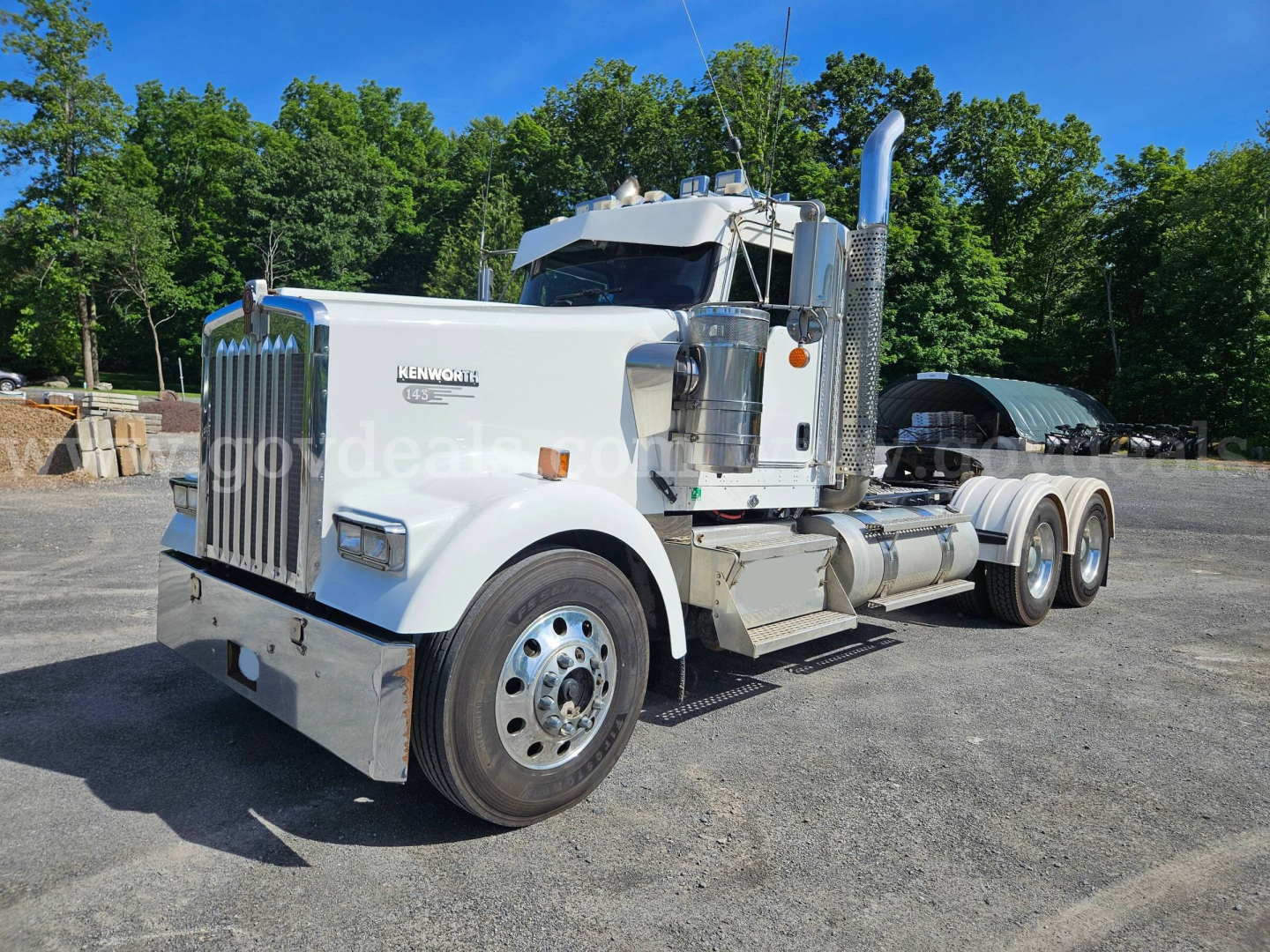 Kenworth W900L heavy truck in front view showing chrome grille, big exhaust stacks, dual rear axles, white cab with Kenworth emblem and chrome bumper.