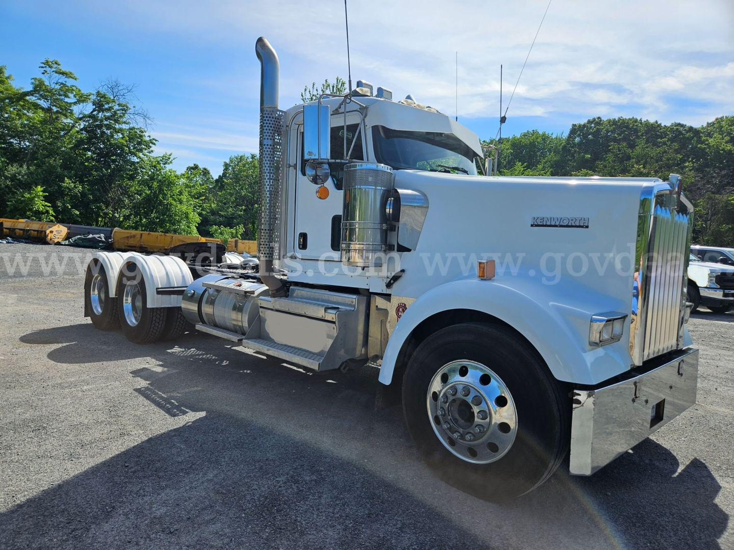 Kenworth W900L truck cab and chassis with chrome exhaust stack, showing Kenworth badge on grille and large front wheel, side fuel tanks, and dual rear axles.