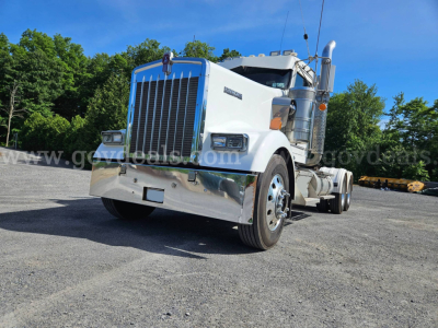Kenworth W900L truck in a yard, front-left view showing large chrome grille, white cab, and dual tires; windshield, mirrors, and exhaust stacks visible.