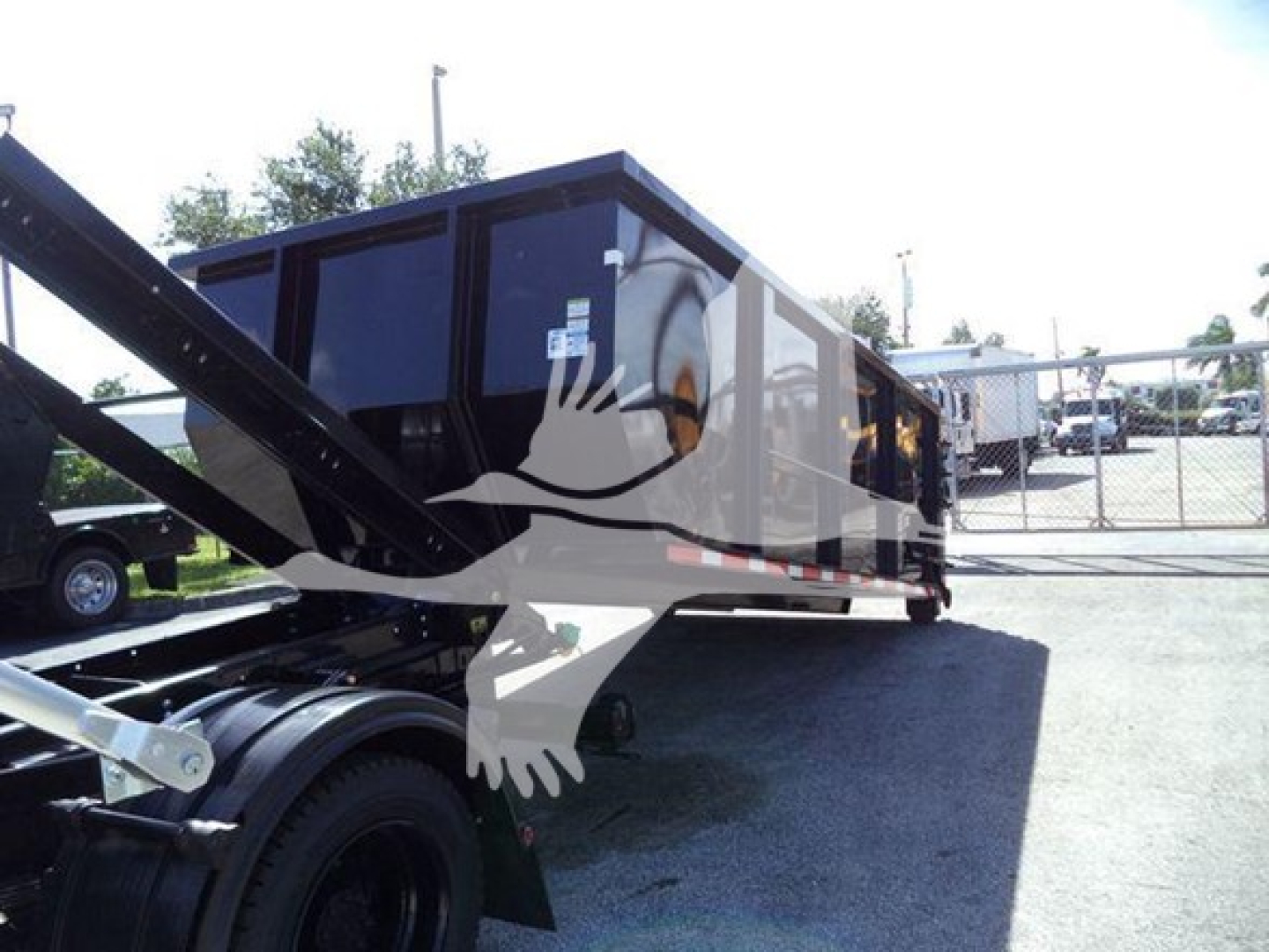 Truck body of a new 2023 Chevrolet Silverado 6500Hd roll-off truck with black cabin, side railing, and behind fence; image shows a portion of the chassis.
