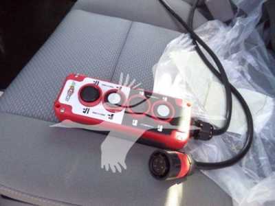 Small red remote control-like device with three circular buttons and a black handle, connected by a cord to a circular connector; sits on a gray vehicle seat.