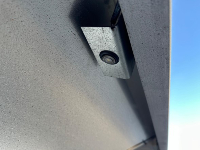 Trailer underside showing a small rectangular sensor/plate with a circular component in the center, mounted to a speckled metal surface.