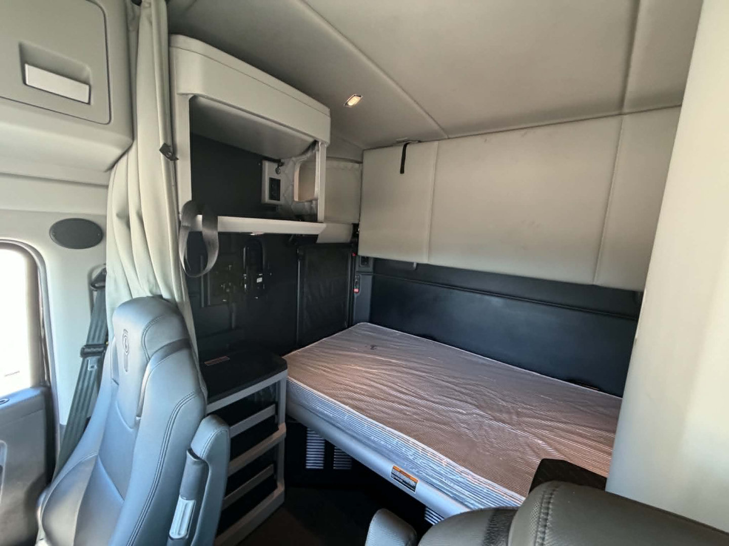 Kenworth T680 sleeper cab showing bunk bed area with overhead storage shelf and light, light gray interior panels.