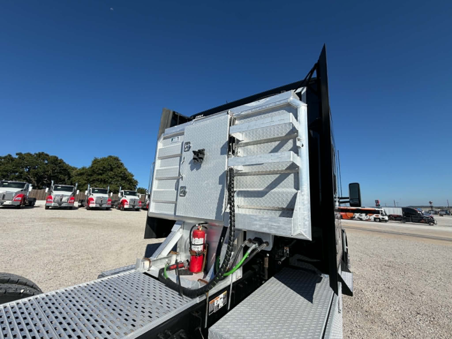 Kenworth T680 truck bed area showing aluminum toolbox with hinged lid, fire extinguisher, and hydraulic lines mounted on a diamond-plate platform; multiple trucks in background.