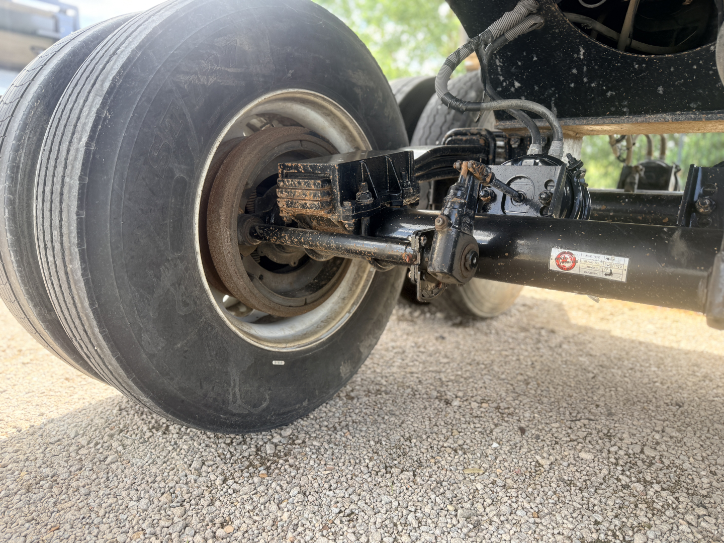 Trailer axle showing a hub with tire and suspension components under a dump trailer. Some labels on the metal frame near the linkage.