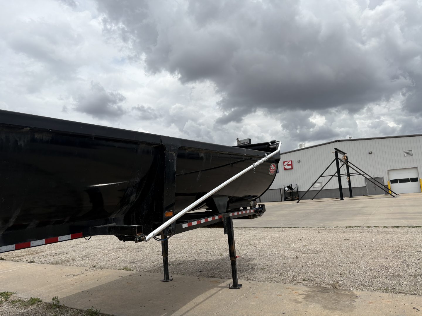 Trailer hitch and support leg of a dump trailer with a black hopper and silver support arm. Ground, warehouse building in background, cloudy sky.
