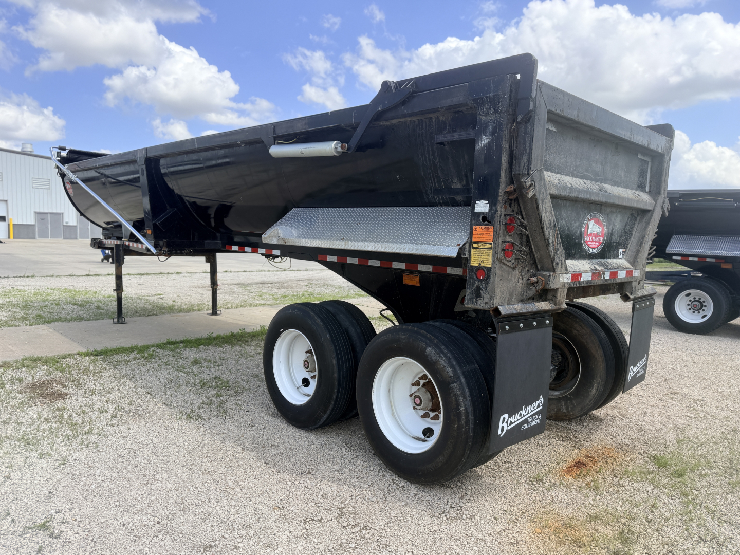 Dump trailer showing dual rear axles with white rims and black dump bed, side marker lights, and brand decals.