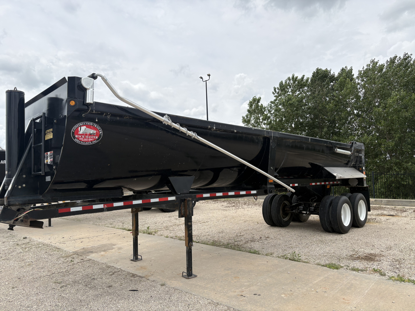 Dump trailer in-service with triple-axle rear, black body and brand sticker on side. Hydraulic beavertail and landing legs visible.
