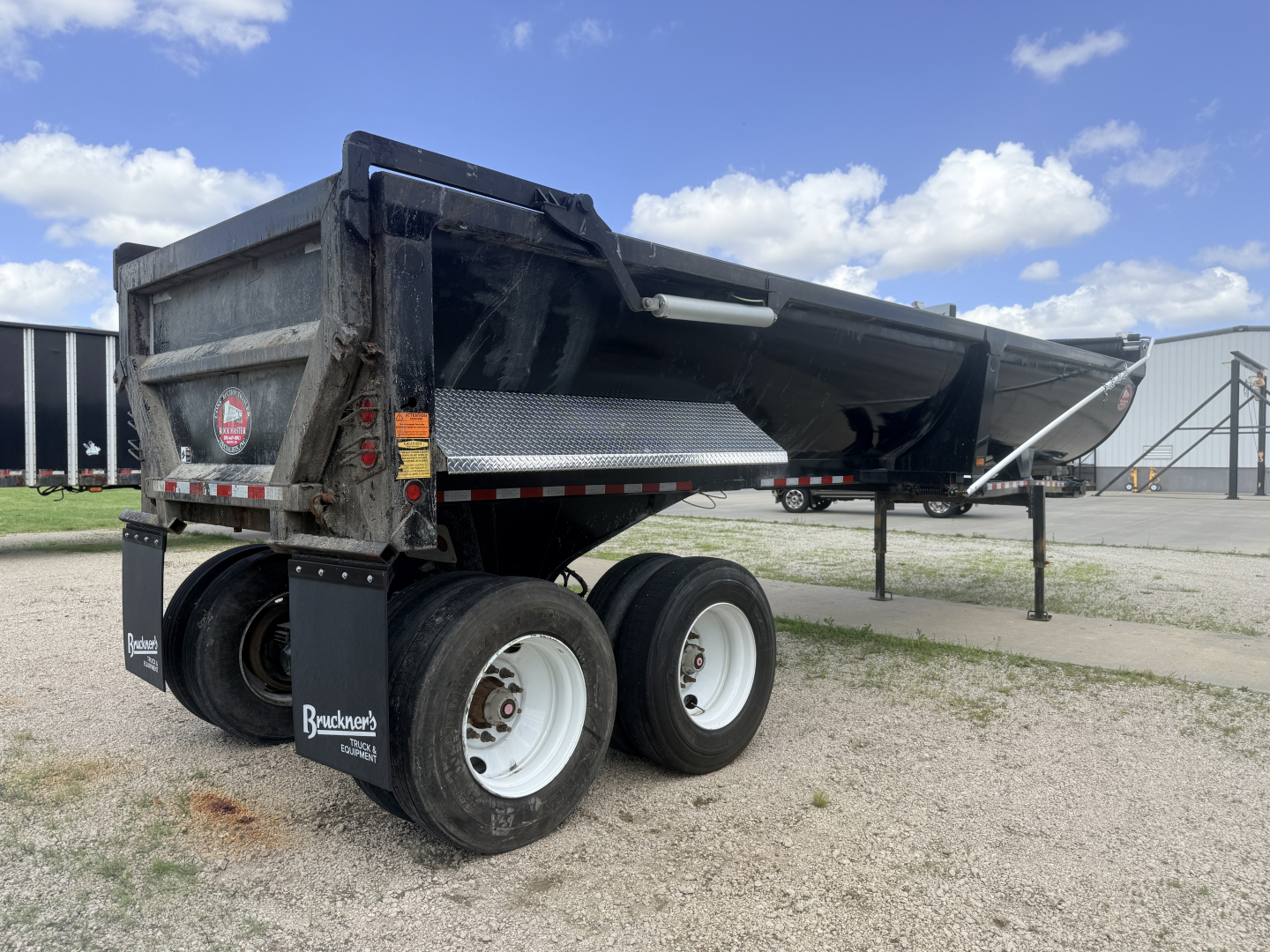 Trailer dump bed with dual rear axles and black metal sides, white wheels, and protective mud flaps. Showing raised bed with Brandenburger Equipment branding on mud flap and reflective stickers.