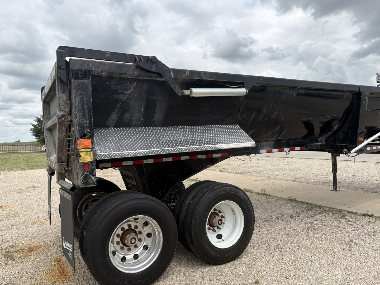 Dump trailer showing dual rear axles with white rims and black metal bed. Side step with diamond plate and white reflective stripe.