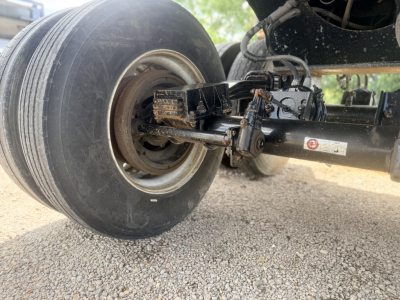 Trailer axle showing a hub with tire and suspension components under a dump trailer. Some labels on the metal frame near the linkage.