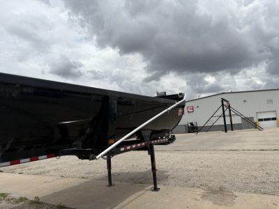 Trailer hitch and support leg of a dump trailer with a black hopper and silver support arm. Ground, warehouse building in background, cloudy sky.