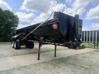 Dump trailer in a parking lot showing a black tandem-axle bed with a raised neck and front stabilizers. Side shows a red circular logo and reflective tape along the crewed edge.