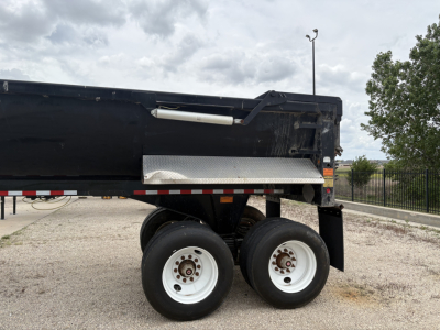 Dump trailer axle view showing dual rear wheels and part of the black dump bed with a metal diamond plate side step.