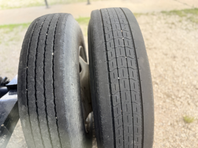 Dual trailer wheels with visible tread patterns; left tire shows wear with small cracks, right tire has more uniform, shallower tread; wheel hubs visible.