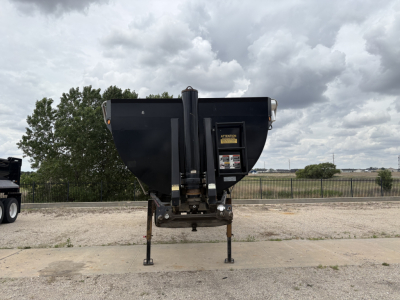 Dump trailer hitch in a raised position with black steel hopper and hydraulic lift, supported by two front legs; rural background and cloudy sky.