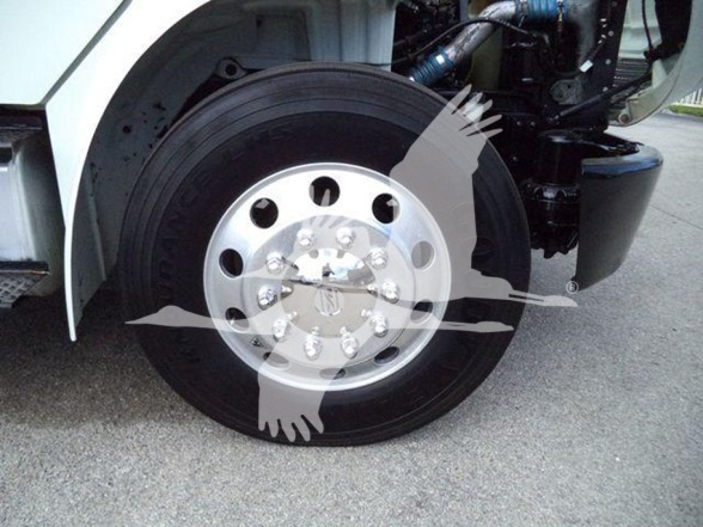 Truck front wheel well showing chrome wheel with multiple lug nuts and part of black tire; part of white truck body and engine components visible.