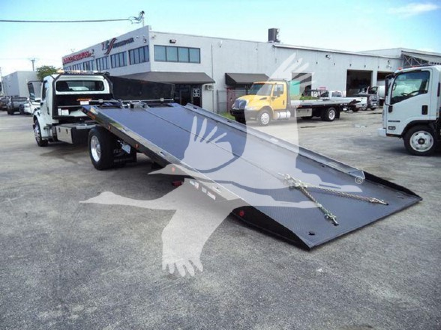 Flatbed tow truck with hydraulic rollback in a truck yard showing Freightliner M2 2019 branding on the bed and surrounding service building.