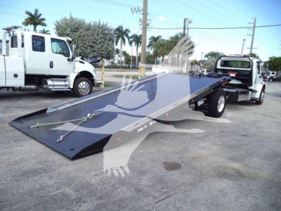Tow truck with flatbed rollback in yard showing winch hooks and ramp surface.