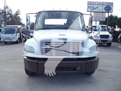 Front view of a white Freightliner M2 106 rollback truck with large grille and bumper. Dashboard text not visible. Signage in background shows 'Truck Sales Center' and 'TIC' logos.