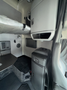 Truck cab interior showing sleeper area and storage cabinet; dashboard area visible near left; vertical console with storage drawers; gray plastic panels and step tread.